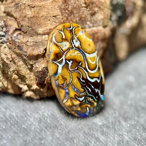 May include: An oval-shaped gemstone with a unique pattern of yellow, brown, white, and blue swirls. The stone is set against a textured brown surface, showcasing its natural beauty. The gemstone's intricate design makes it a striking piece.
