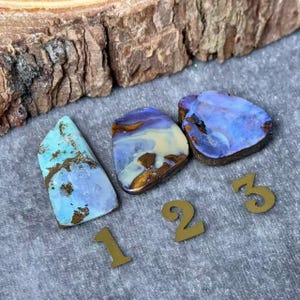 Puede incluir: Tres piedras preciosas de ópalo pulidas en tonos azules, morados y dorados. Cada piedra tiene un patrón único y está numerada con números de metal dorado. Las piedras preciosas se exhiben sobre una superficie gris con un fondo de madera.