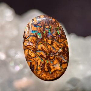 Peut inclure: Une pierre précieuse d'opale de forme ovale avec un motif unique de tourbillons bruns, oranges et blancs. L'opale présente des éclairs d'iridescence bleue et verte, créant un effet visuel saisissant. La pierre est polie.