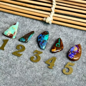 May include: Five polished opal gemstones in various shapes and colours, including teal, blue, and brown, are displayed with brass number markers. The opals are arranged on a grey surface with a bamboo element in the background.