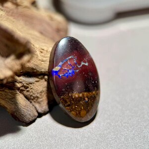 Peut inclure: Pierre précieuse ovale avec une base rouge foncé, présentant des motifs bleus et blancs. La partie inférieure a un aspect brun et texturé. La pierre est posée sur un fond en bois flou.