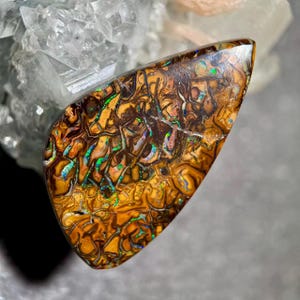 Peut inclure: Une pierre précieuse polie, de forme libre, avec un motif complexe de teintes marron, dorées et vertes et bleues irisées. La pierre a une apparence unique et organique, avec un réseau de lignes et de textures sur sa surface. Il s'agit probablement d'un type d'opale.