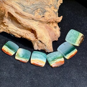 May include: Seven square-shaped gemstones with a layered appearance. The stones feature bands of green, blue, orange, and brown hues. A piece of textured, light brown wood sits in the background on a black surface.