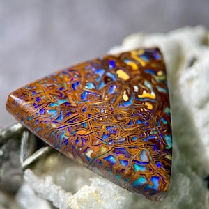 May include: A triangular-shaped gemstone with a complex pattern of brown, blue, yellow, and teal colors. The stone has a polished surface and a unique, organic appearance. The gemstone is likely a type of opal, suitable for jewelry or collection.