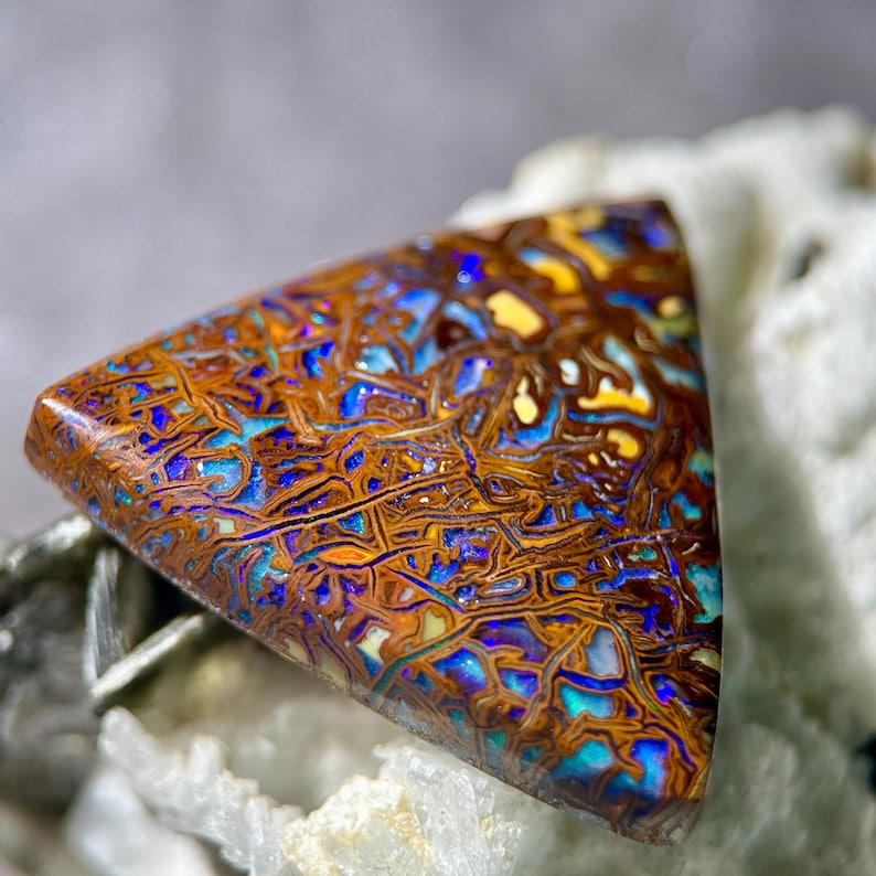 May include: A triangular-shaped gemstone with a complex pattern of brown, blue, yellow, and teal colors. The stone has a polished surface and a unique, organic appearance. The gemstone is likely a type of opal, suitable for jewelry or collection.