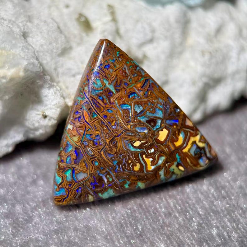 May include: A triangular gemstone with a mosaic pattern of brown, blue, yellow, and orange hues. The polished surface of the stone reflects light, showcasing the intricate internal structure. The gemstone is likely a type of opal.