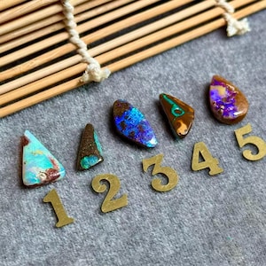 May include: Polished opal gemstones in diverse shapes and colours, including blue, green, and purple. The gemstones are arranged with small brass numbers on a grey surface, with a wooden bamboo blind in the background.