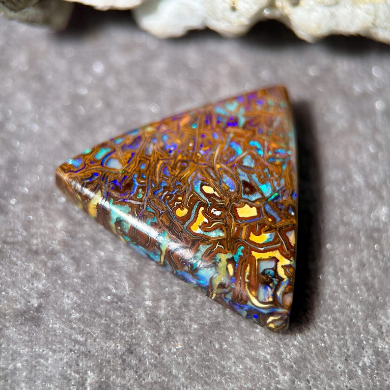 May include: A triangular-shaped gemstone with a mosaic pattern of brown, yellow, blue, and green hues. The stone has a polished surface and a natural, organic appearance. The gemstone is likely a type of opal.
