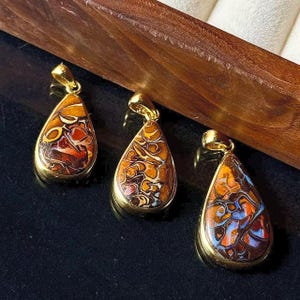 May include: Three teardrop-shaped pendants with gold settings. The gemstones display intricate patterns in brown, orange, and red. The pendants are presented on a dark surface, with a wooden element in the background.