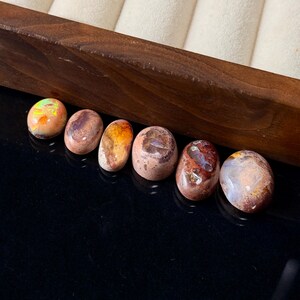 May include: A collection of six oval-shaped opals, each displaying unique patterns and colors. The opals range in hues from orange and brown to iridescent flashes of green and yellow. They are arranged on a black surface next to a wooden box.