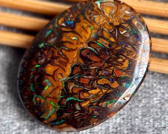 May include: Oval-shaped opal gemstone with a polished surface. The stone features a swirling pattern of brown, orange, and white hues, with flashes of green and blue iridescence. The gemstone is set against a neutral background.