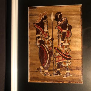 Può includere: Un'opera d'arte incorniciata che raffigura due figure in abiti tradizionali africani. Le figure sono realizzate in paglia intrecciata e sono posizionate su uno sfondo marrone. L'opera d'arte è incorniciata in nero.