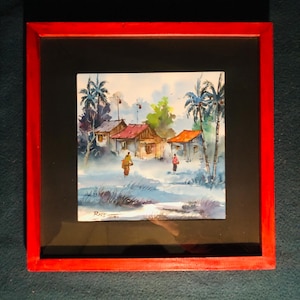 Peut inclure: Une peinture à l'aquarelle encadrée représentant une scène de village rural avec des maisons, des palmiers et des personnages marchant le long d'un chemin. La peinture est signée "Ries 2018" dans le coin inférieur gauche.
