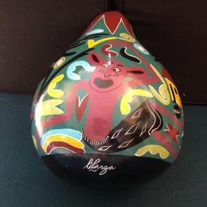 Pode incluir: Uma cabaça em forma de pêra pintada com um design caprichoso que apresenta uma criatura vermelha com uma boca grande e olhos negros. A cabaça é pintada em tons de verde, vermelho, amarelo, preto e branco. A cabaça é assinada "libanga" na parte inferior.
