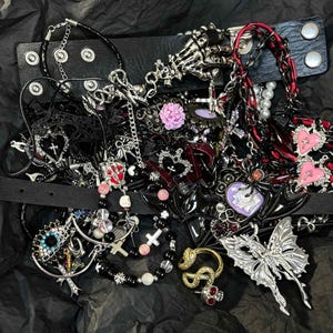Puede incluir: Una colección de joyas de temática gótica, que incluye collares, pulseras y gargantillas. Las piezas presentan cruces, corazones, calaveras y diseños de mariposas. La paleta de colores incluye acentos negros, plateados, rojos y rosas. Las joyas están dispuestas sobre una superficie negra.