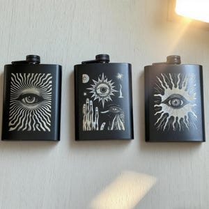 May include: Three black flasks with unique designs. The flasks feature eye and sunburst illustrations in white. Each flask has a black cap. The flasks are rectangular and appear to be made of metal.