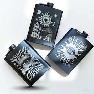 May include: Three black flasks with silver-colored, eye-themed illustrations. Each flask has a different design, including an eye with radiating lines, an eye within a sunburst, and a design with an eye, hand, and other celestial elements. The flasks have a matte finish.