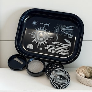 May include: A black metal rolling tray with a rounded rectangular shape, featuring a white graphic design of an eye, hand, and sun. Includes a matching grinder and accessories. The tray is approximately 20 cm long.