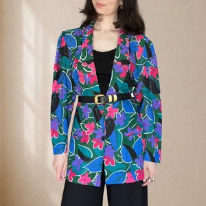 Könnte beinhalten: Ein langärmeliger Blazer mit einem lebendigen Blumenmuster in den Farben Pink, Violett und Blau auf dunkelgrünem Grund. Der Blazer verfügt über ein steigendes Revers, einen Knopfverschluss auf der Vorderseite und wird mit einem schwarzen Gürtel.
