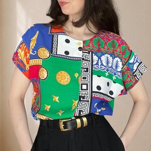 Könnte beinhalten: Eine kurzärmelige Bluse mit einem lebendigen Patchwork-Design in kräftigen Blöcken aus Grün, Blau, Rot und Weiß. Das Muster zeigt goldene kreisförmige Medaillons, schwarze Punkte und komplexe geometrische griechische Mäanderbordüren auf Stoff.