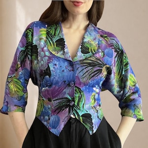 Könnte beinhalten: Ein kurz geschnittener Blazer mit einem lebendigen, malerischen Druck aus violetten Trauben und grünen Blättern auf blauem Grund. Das Kleidungsstück hat Dreiviertelärmel, einen Reverskragen und eine Knopfleiste vorne mit zwei passenden Knöpfen.