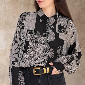 Op de afbeelding: Een overhemd met lange mouwen en knoopsluiting met een zwart-wit paisley patchworkpatroon. Het overhemd heeft een klassieke kraag, zwarte knopen en losvallende mouwen. Het is ingestopt in een zwarte broek met een riem met een gouden gesp.