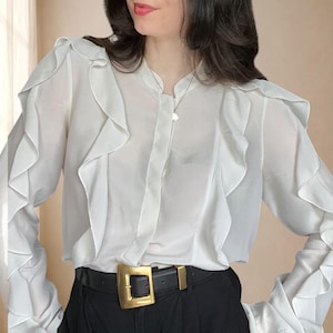 Könnte beinhalten: Eine weiße Langarmbluse mit Stehkragen und vertikalen Rüschen, die von den Schultern nach unten verlaufen. Die Bluse ist in eine schwarze Hose gesteckt, die mit einem schwarzen Ledergürtel mit großer goldfarbener Schnalle gehalten wird.