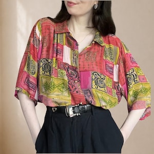 Op de afbeelding: Een overhemd met korte mouwen en knoopsluiting met een levendig patchworkpatroon in tinten rood, roze, geel en groen met zwarte decoratieve motieven. Het overhemd is in een zwarte broek gestoken, vastgezet met een zwarte leren riem met een zilveren gesp.