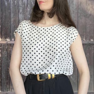 Könnte beinhalten: Eine weiße kurzärmelige Bluse mit schwarzem Punktemuster, kombiniert mit einer schwarzen Hose, die von einem schwarzen Ledergürtel mit goldfarbener Metallschnalle gehalten wird. Die Bluse hat einen Rundhalsausschnitt und eine lockere Passform.