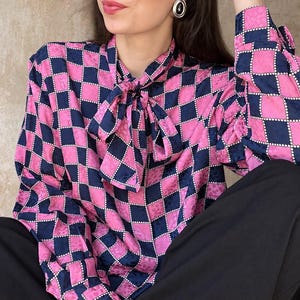 Könnte beinhalten: Eine langärmelige Bluse mit einem lebhaften pinkfarbenen und marineblauen Karomuster mit einer dezenten Ton-in-Ton-Textur. Das Kleidungsstück verfügt über einen hohen Ausschnitt mit einem integrierten Bindedetail und langen Ärmeln mit Manschetten.