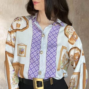 May include: A long-sleeved button-down shirt featuring a white base with a central vertical purple panel containing a diamond pattern. The shirt is decorated with various framed illustrations of cats and floral arrangements in brown and gold tones.