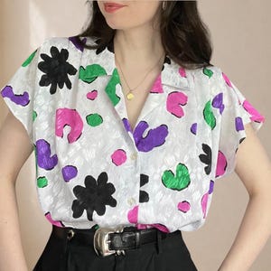 Könnte beinhalten: Ein weißes, kurzärmeliges Hemd mit Knopfleiste aus strukturiertem Stoff, bedruckt mit einem bunten, abstrakten Muster aus schwarzen, leuchtend pinken, violetten und grünen Formen. Das Hemd hat einen Reverskragen und steckt in einer schwarzen Hose.