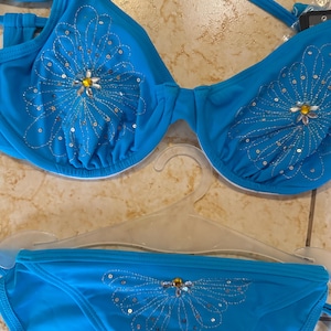 Peut inclure: Ensemble de bikini turquoise avec un motif floral. Le haut et le bas sont ornés d'un motif de fleur à paillettes, centré d'un bijou jaune. Le bikini est présenté sur un cintre transparent.