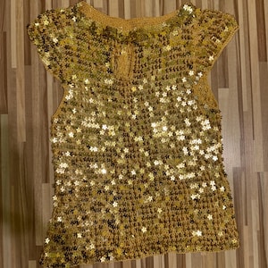 Puede incluir: Una blusa de crochet dorada, con cuello redondo y mangas cortas. La blusa está adornada con pequeños detalles brillantes, creando un efecto texturizado y brillante. La prenda tiene un detalle de ojo de cerradura en la espalda.