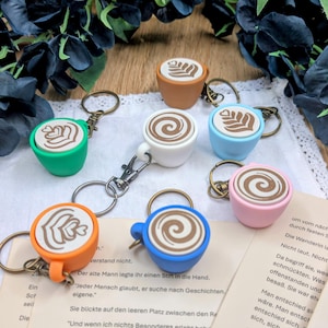 Könnte beinhalten: Eine Sammlung von sieben Miniatur-Kaffeetassen-Schlüsselanhängern in verschiedenen Farben wie Grün, Orange, Weiß, Blau, Hellblau, Rosa und Braun. Jede Tasse zeigt ein detailliertes Latte-Art-Design auf der Oberfläche und hat einen Metallring.
