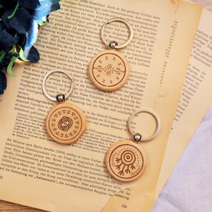 Könnte beinhalten: Drei runde Holzschlüsselanhänger mit silbernen Metallringen. Jeder Schlüsselanhänger hat ein einzigartiges lasergraviertes Design: eines mit einer zentralen Spirale und Kreuzen, eines mit einem zentralen Knotensymbol und eines mit einem Zielmuster.