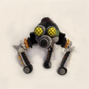 May include: A soft toy depicting a robotic creature. The toy is mainly black and grey, with yellow detailing on the eyes and orange accents. It has three legs and a circular body.