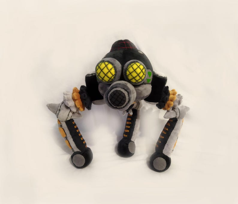May include: A soft toy depicting a robotic creature. The toy is mainly black and grey, with yellow detailing on the eyes and orange accents. It has three legs and a circular body.