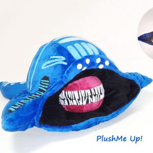 May include: A blue plush toy creature with a large open mouth, revealing a pink interior and white teeth. A matching illustration is on a white background. The text "PlushMe Up!" is at the bottom of the image.