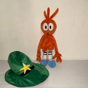May include: An orange plush toy character with a green hat featuring a yellow star and black band. The character has white eyes, a black mouth, and blue shoes. The hat is next to the character.
