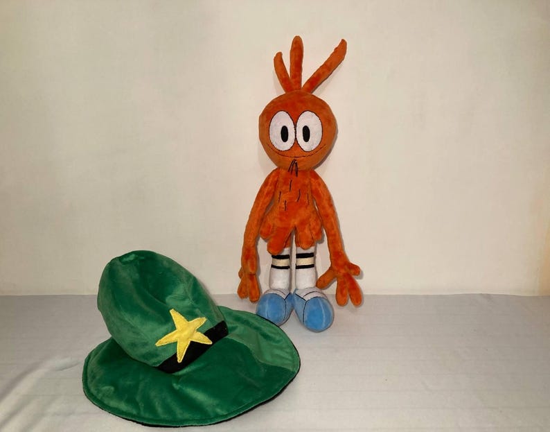 May include: An orange plush toy character with a green hat featuring a yellow star and black band. The character has white eyes, a black mouth, and blue shoes. The hat is next to the character.