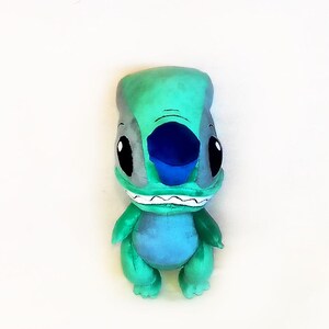 Peut inclure: Peluche du personnage Stitch du film Lilo & Stitch. La peluche est principalement turquoise avec des accents gris, un nez bleu et de grands yeux noirs. Il a une expression souriante avec des dents blanches.