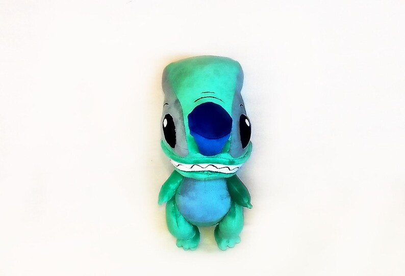 Peut inclure: Peluche du personnage Stitch du film Lilo & Stitch. La peluche est principalement turquoise avec des accents gris, un nez bleu et de grands yeux noirs. Il a une expression souriante avec des dents blanches.