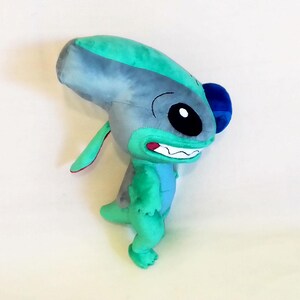Peut inclure: Une peluche repr&eacute;sentant une cr&eacute;ature extraterrestre bleue et verte avec de grandes oreilles et un large sourire. Le jouet a une grosse t&ecirc;te, de petits bras et un petit corps. La cr&eacute;ature est sur une surface blanche.