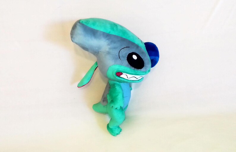 Peut inclure: Une peluche repr&eacute;sentant une cr&eacute;ature extraterrestre bleue et verte avec de grandes oreilles et un large sourire. Le jouet a une grosse t&ecirc;te, de petits bras et un petit corps. La cr&eacute;ature est sur une surface blanche.