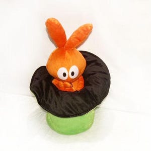 May include: A plush orange bunny toy with big eyes emerges from a black top hat. The hat rests on a green cylinder base. The toy is set against a white background.