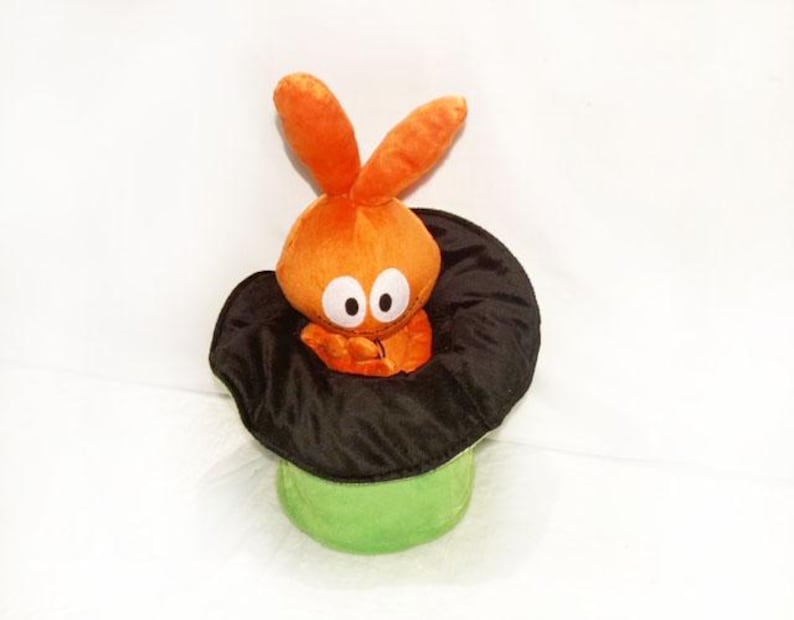 May include: A plush orange bunny toy with big eyes emerges from a black top hat. The hat rests on a green cylinder base. The toy is set against a white background.