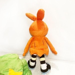 May include: A plush toy bunny in orange, featuring long ears, arms, and legs. The bunny wears black and white striped socks and black shoes with blue soles. A green plush object with a yellow star is partially visible.