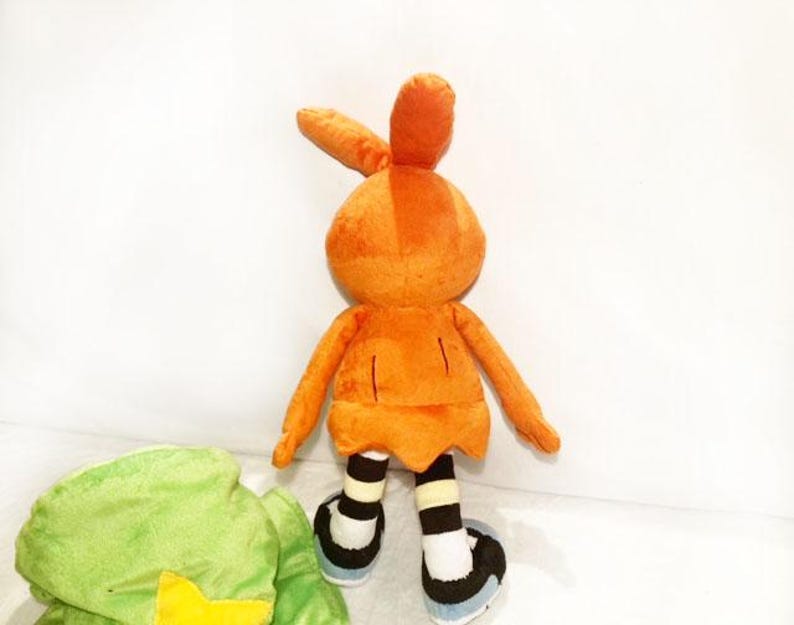 May include: A plush toy bunny in orange, featuring long ears, arms, and legs. The bunny wears black and white striped socks and black shoes with blue soles. A green plush object with a yellow star is partially visible.