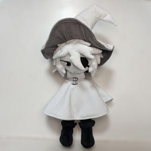 K&ouml;nnte beinhalten: Pl&uuml;schpuppe einer Figur mit wei&szlig;em Kleid, schwarzen Stiefeln und einem grau-wei&szlig;en Hexenhut. Die Puppe hat wei&szlig;es Haar und eine schwarze Augenklappe. Die Puppe steht aufrecht vor einem wei&szlig;en Hintergrund.